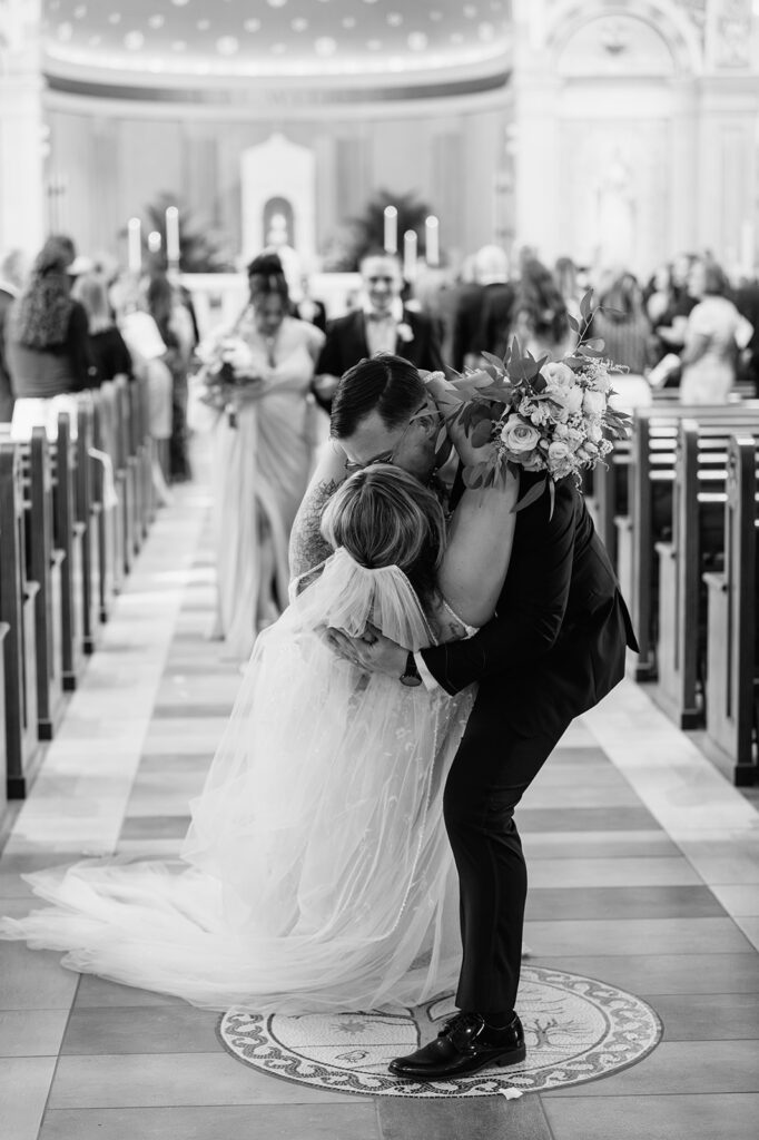 Black and white photo of a bride and groom kissing at the end of the aisle during their wedding ceremony at St. Pius X Catholic Church