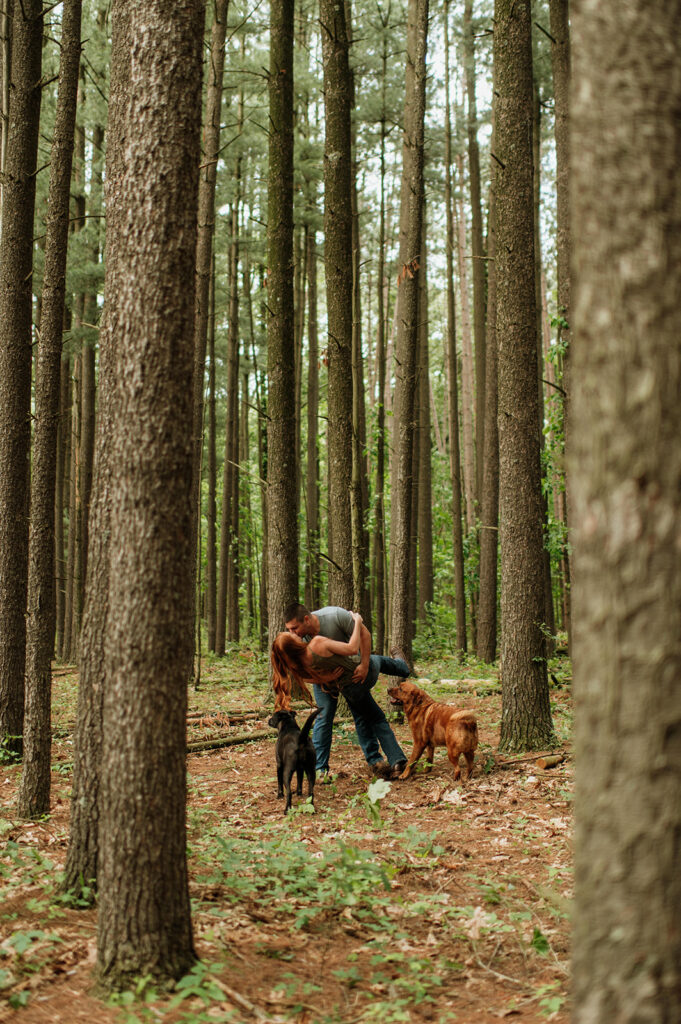 Man kissing his fiancé with their two dogs in the woods