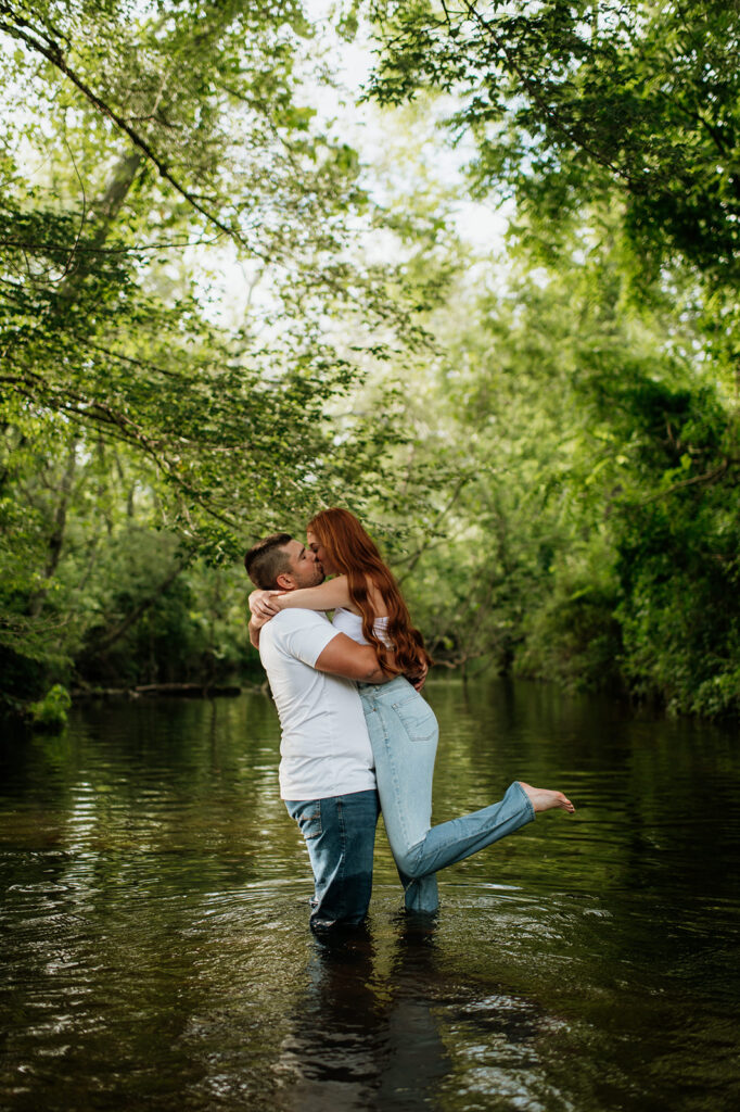 Man lifting woman up in a creek for their outdoor summer engagement photos in Indiana