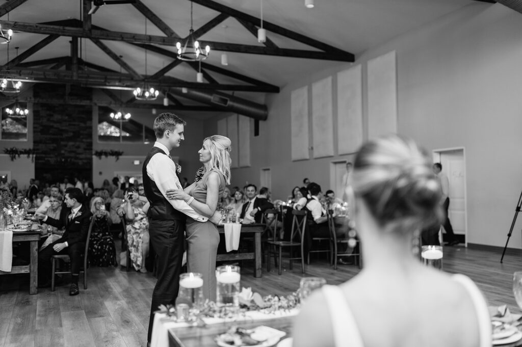 Black and white photo of a groom dancing with his mother as the bride watches from afar