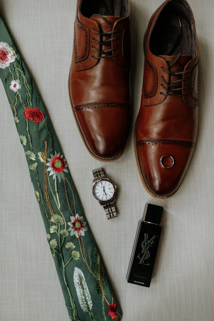 Grooms wedding details with shoes, cologne, a watch, and a green wildflower tie