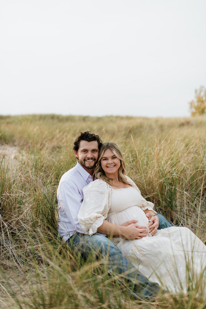 Man and woman sitting in beach grass at New Buffalo Beach for their maternity photos in Michigan