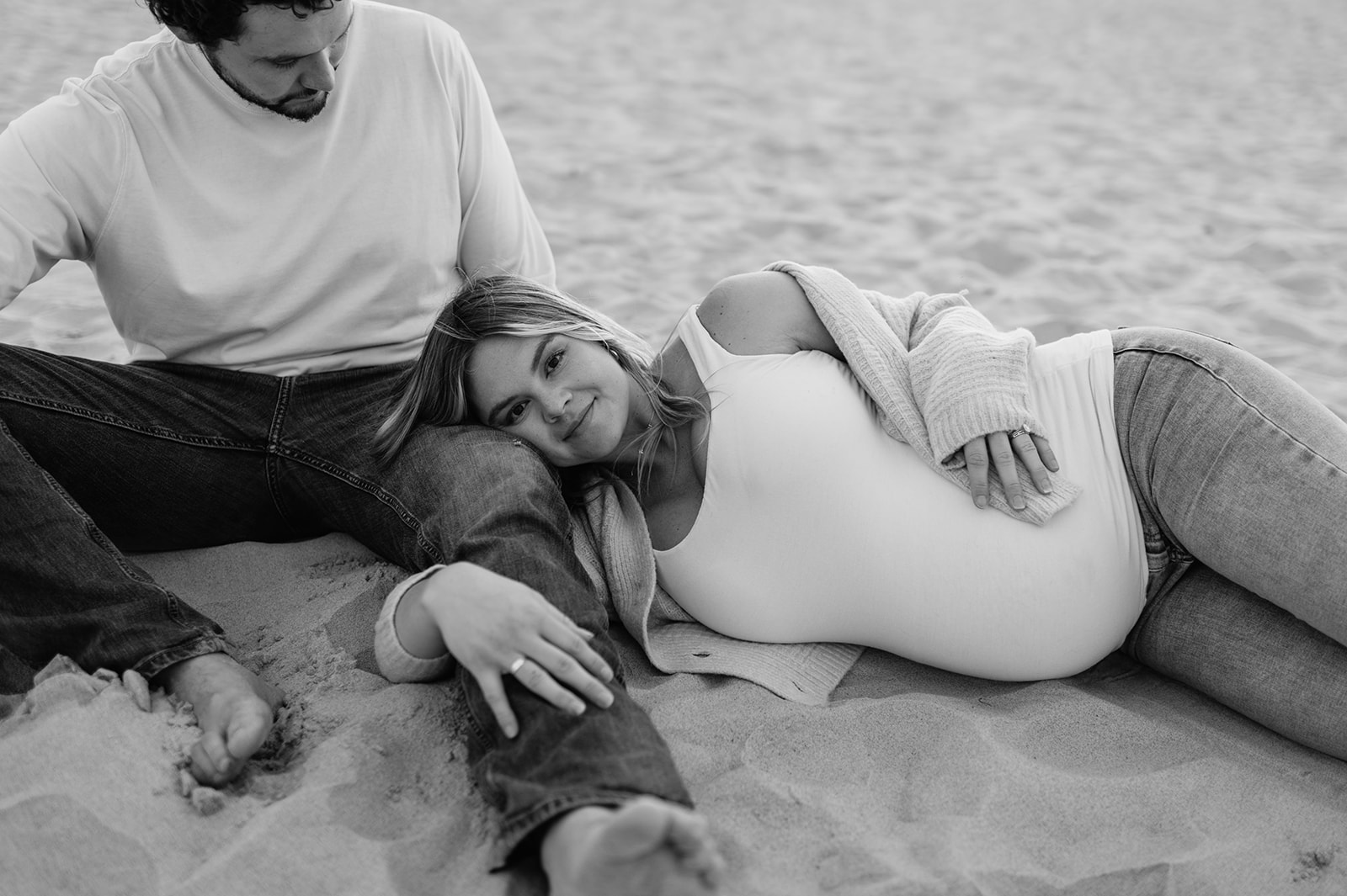 Black and white photo of a pregnant woman laying on her husband on the beach