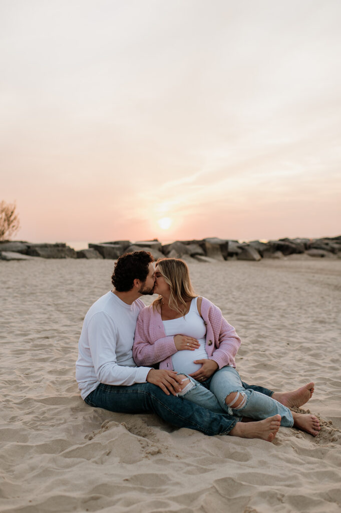 Couple kissing as they sit on the beach during their Michigan maternity photos