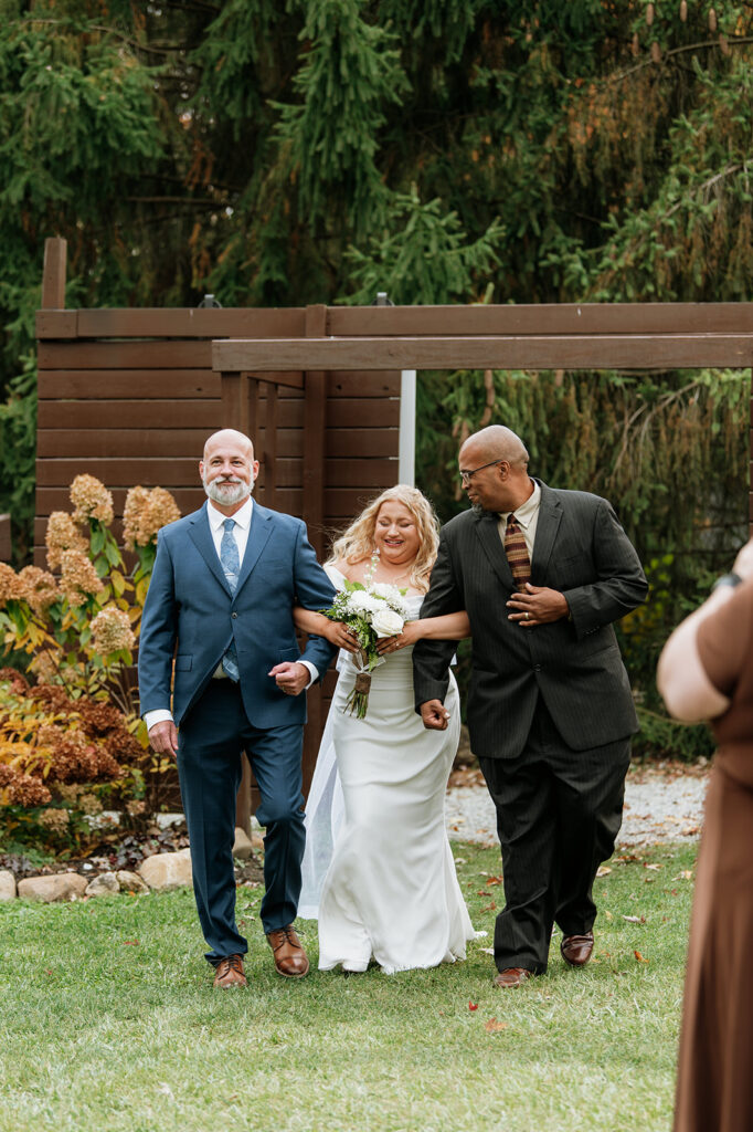 Bride being walked down the aisle by her father and stepfather for her outdoor fall Willow Acres wedding ceremony on the Pergola