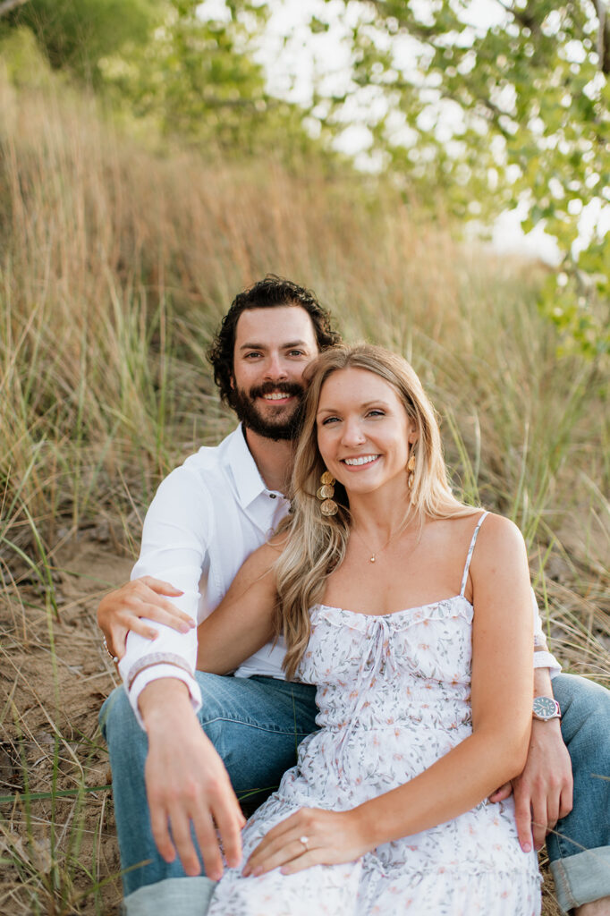 Couple snuggling up while sitting in the beach grass at Indiana Dunes State Park for their outdoor summer engagement photos