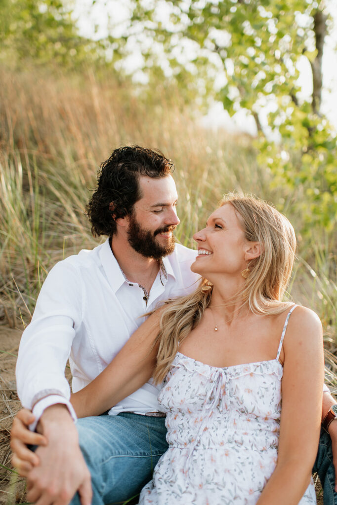 Couple snuggling up while sitting in the beach grass at Indiana Dunes State Park for their outdoor summer engagement photos