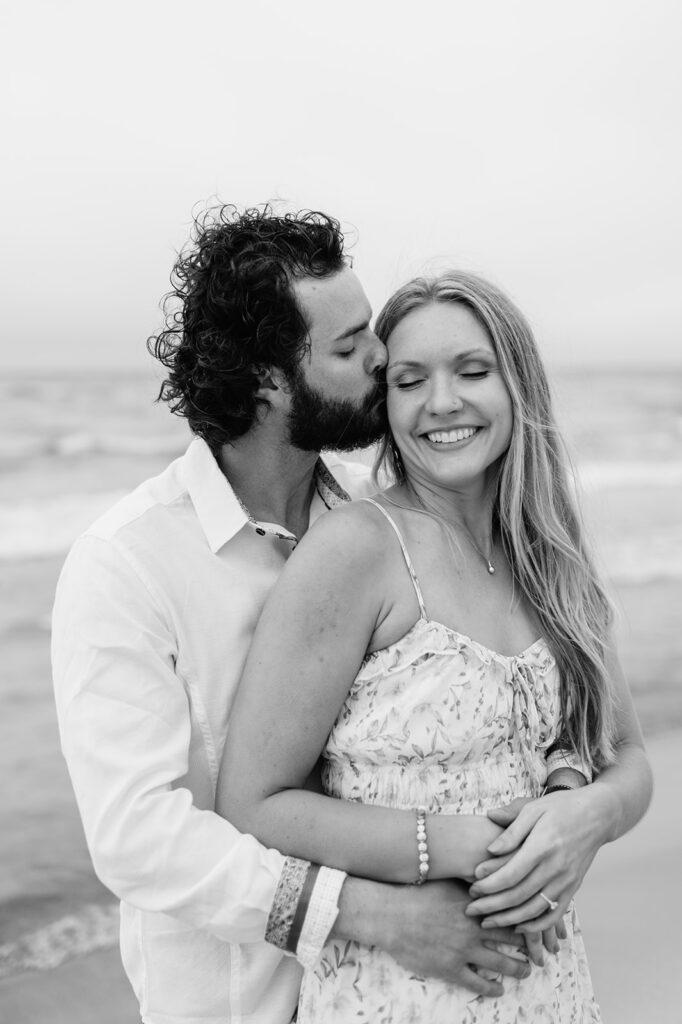Black and white photo of a man kissing his fiancé during their beach engagement session