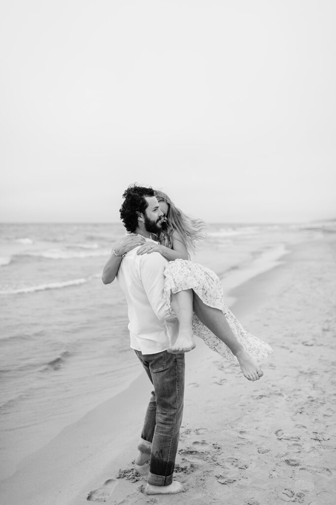 Black and white photo of a man picking up his fiancé during their playful beach engagement session