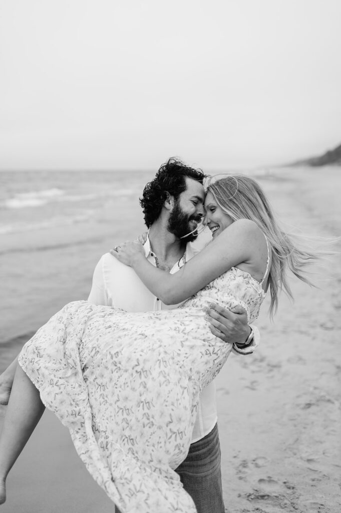 Black and white photo of a man picking up his fiancé during their playful beach engagement session