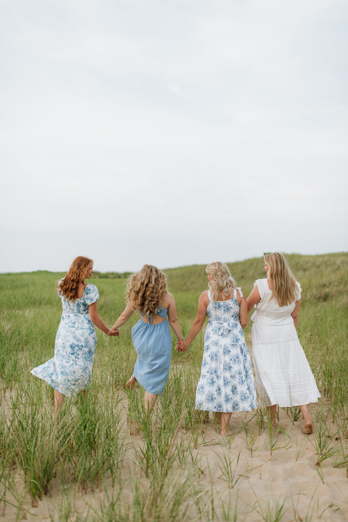 Four sisters holding hands and walking in the beach grass during their outdoor extended family photoshoot on New Buffalo Beach in Michigan