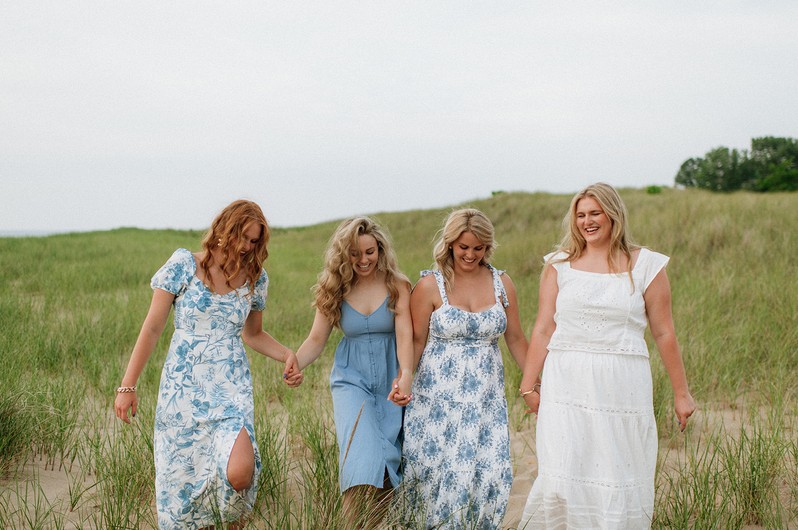 Four sisters holding hands and walking in the beach grass during their outdoor extended family photoshoot on New Buffalo Beach in Michigan
