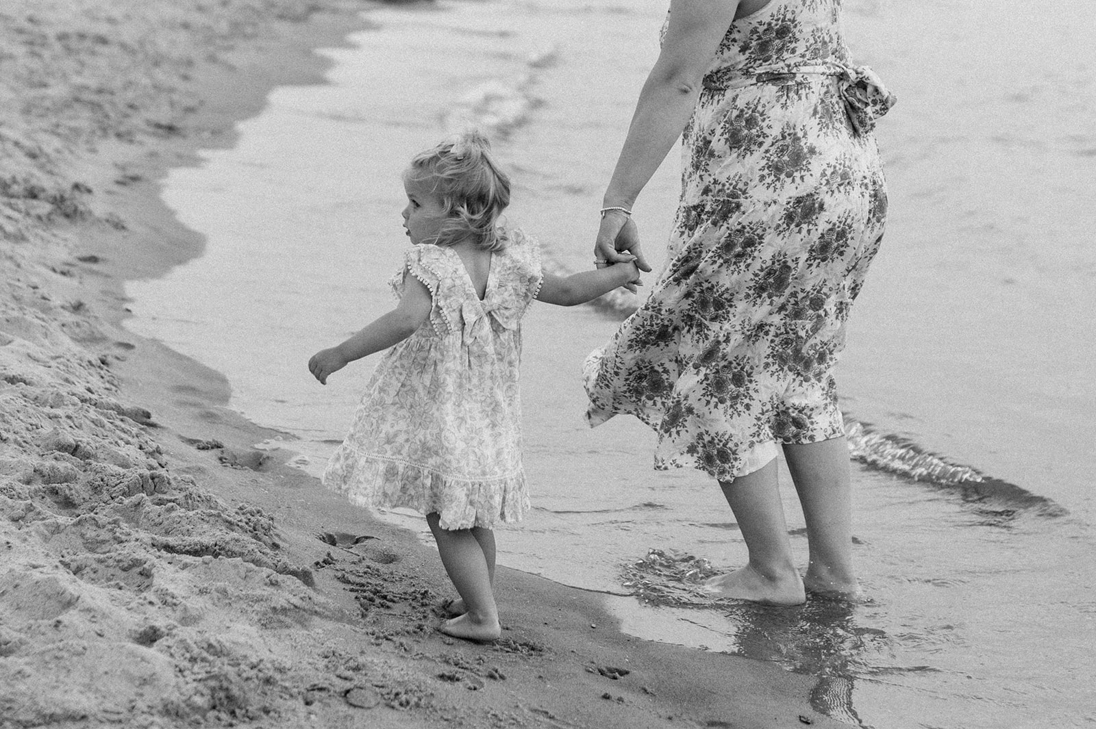 Close up black and white photo of a toddler holding her mothers hand as they walk the shoreline