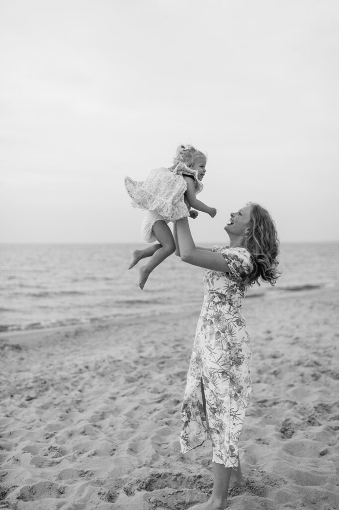 Black and white photo of an aunt throwing her niece in the air during their extended family photoshoot at New Buffalo Beach in Michigan
