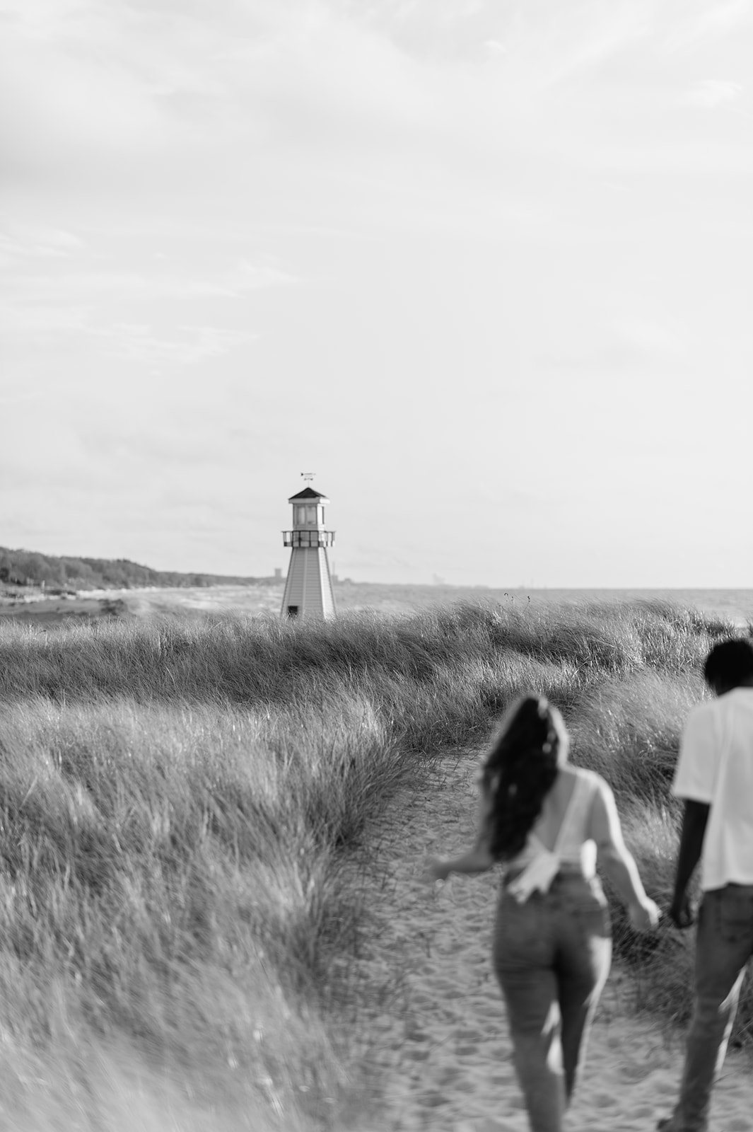 Black and white photo of a couple walking the trails for their New Buffalo Michigan engagement photos with a lighthouse in the background