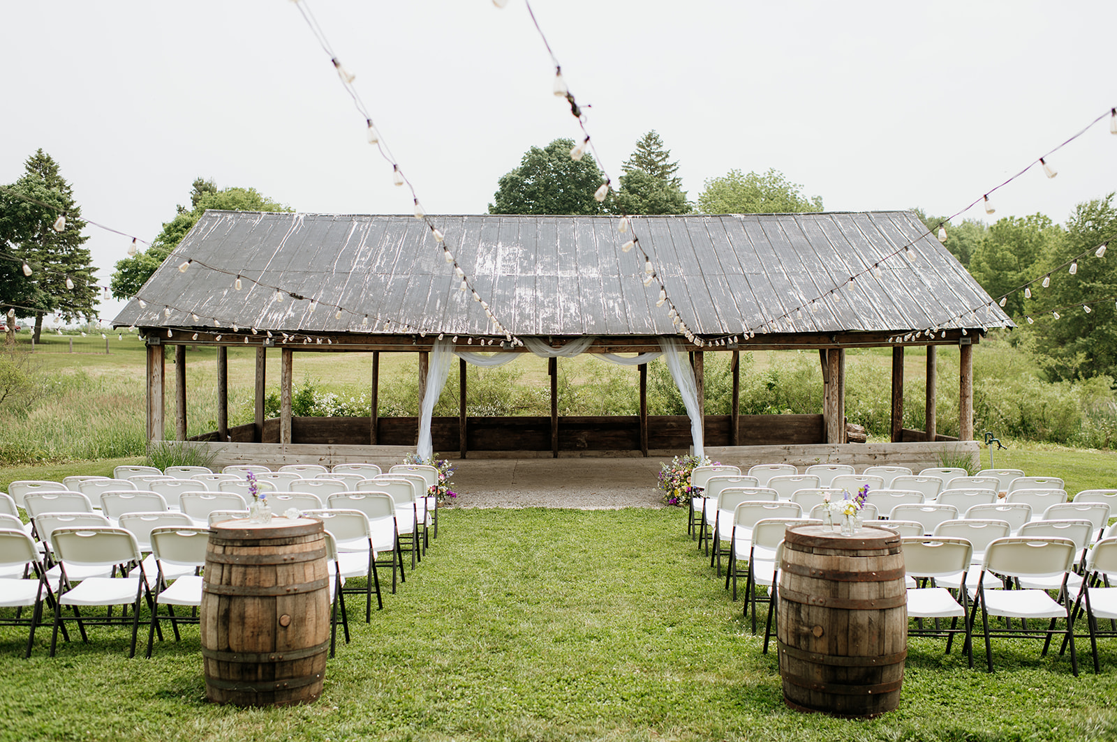 Wide shot of the outdoor ceremony space at 5C Farms with rows of white chairs, string lights, and rustic wood shelter decorated with draped white fabric