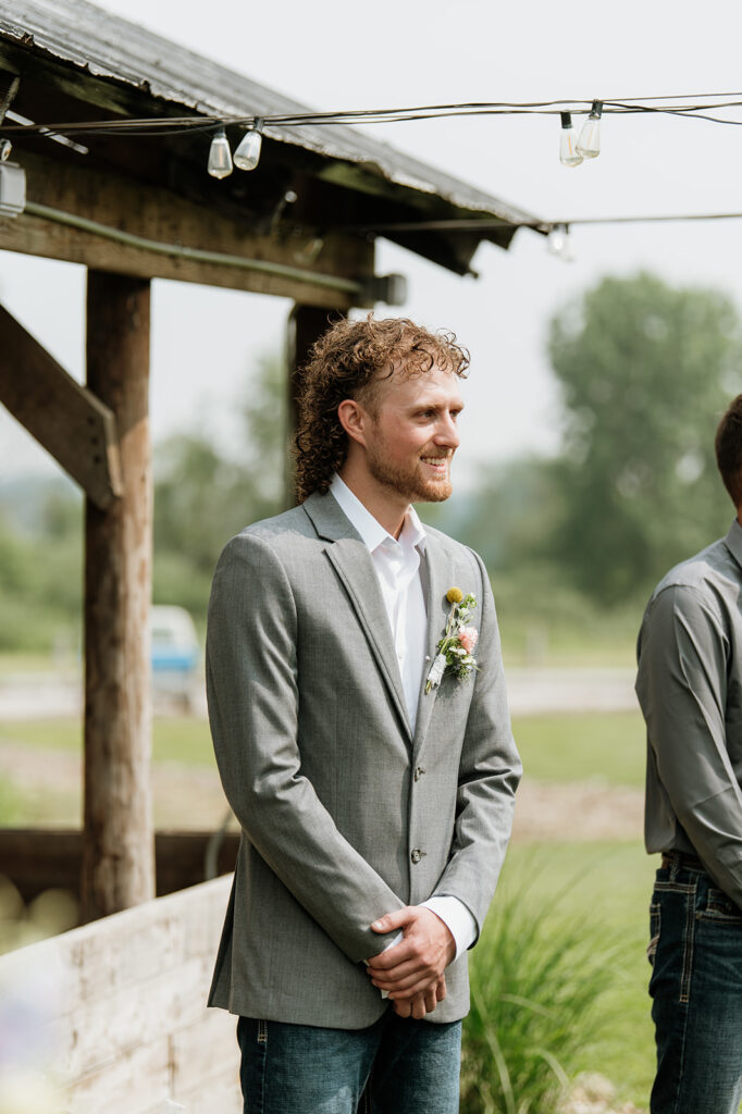 Groom in gray sport coat, jeans, and boots smiling as he waits at the altar during Indiana barn wedding ceremony.