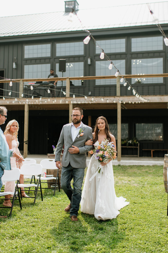 Bride walking down the aisle with her father, carrying a colorful bouquet, with the black barn at 5C Farms in the background.