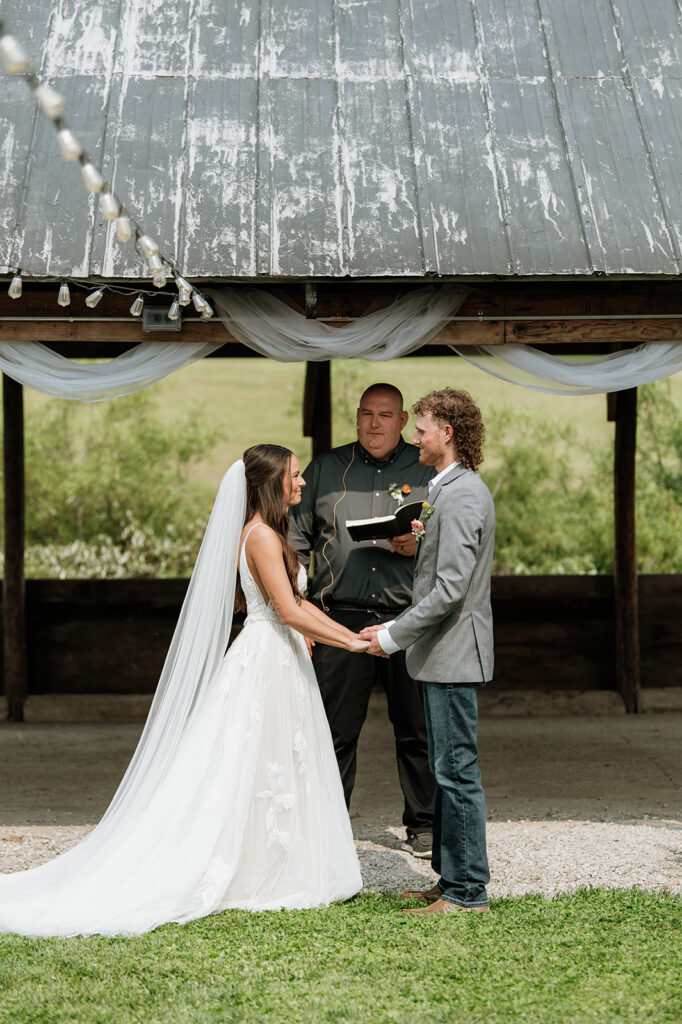 Bride and groom holding hands under a rustic wooden shelter draped with white fabric as they exchange vows.