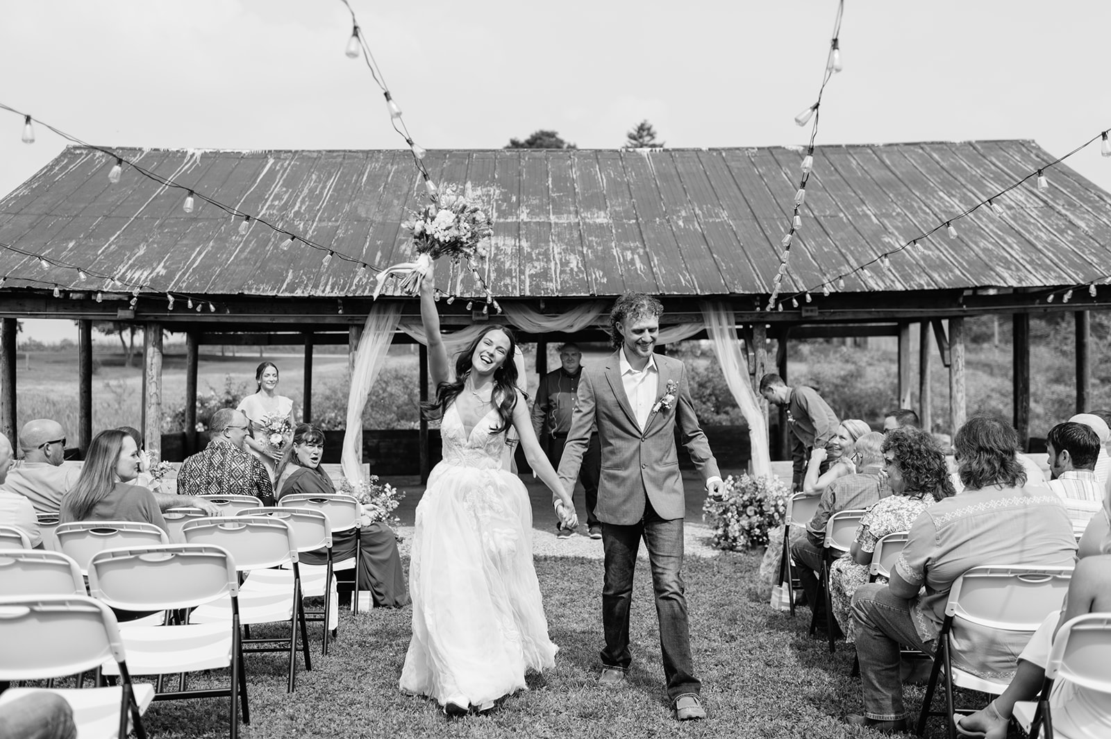 Black-and-white photo of the bride and groom walking back up the aisle together, the bride raising her bouquet in celebration.