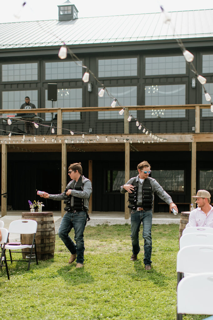 Two groomsmen dressed casually in jeans and vests act as “beer boys,” handing out drinks to guests before the ceremony.