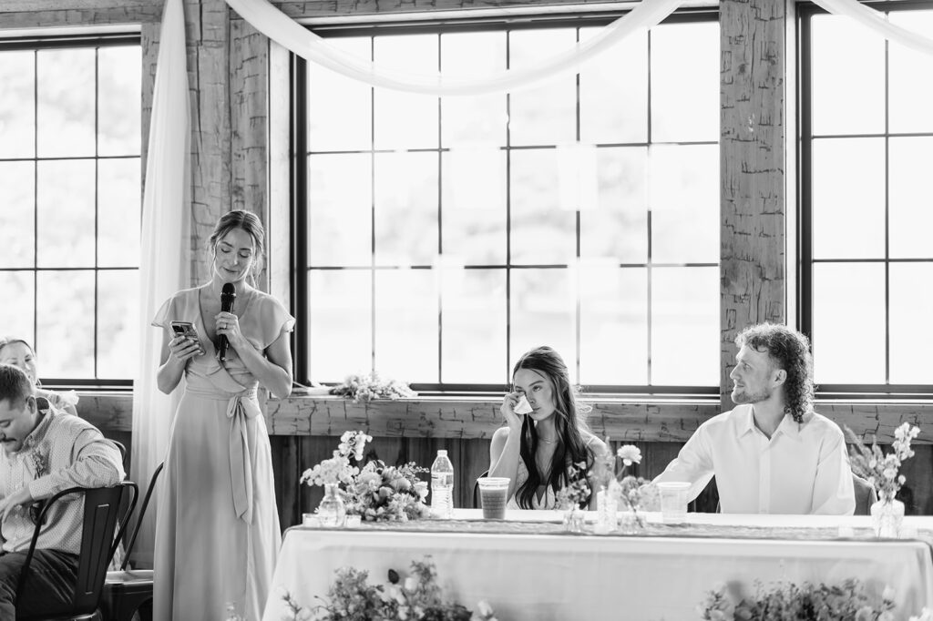 Black-and-white photo of maid of honor giving a toast while the bride wipes away a tear, with the groom seated beside her.