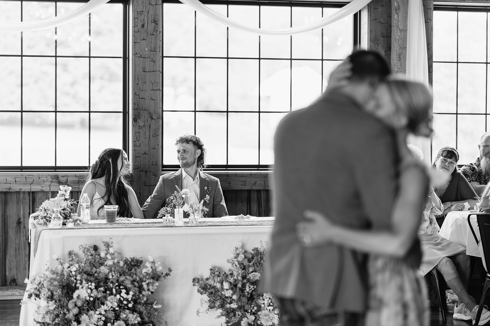 Black-and-white photo of bride and groom smiling at each other while while the brides parents share a dance