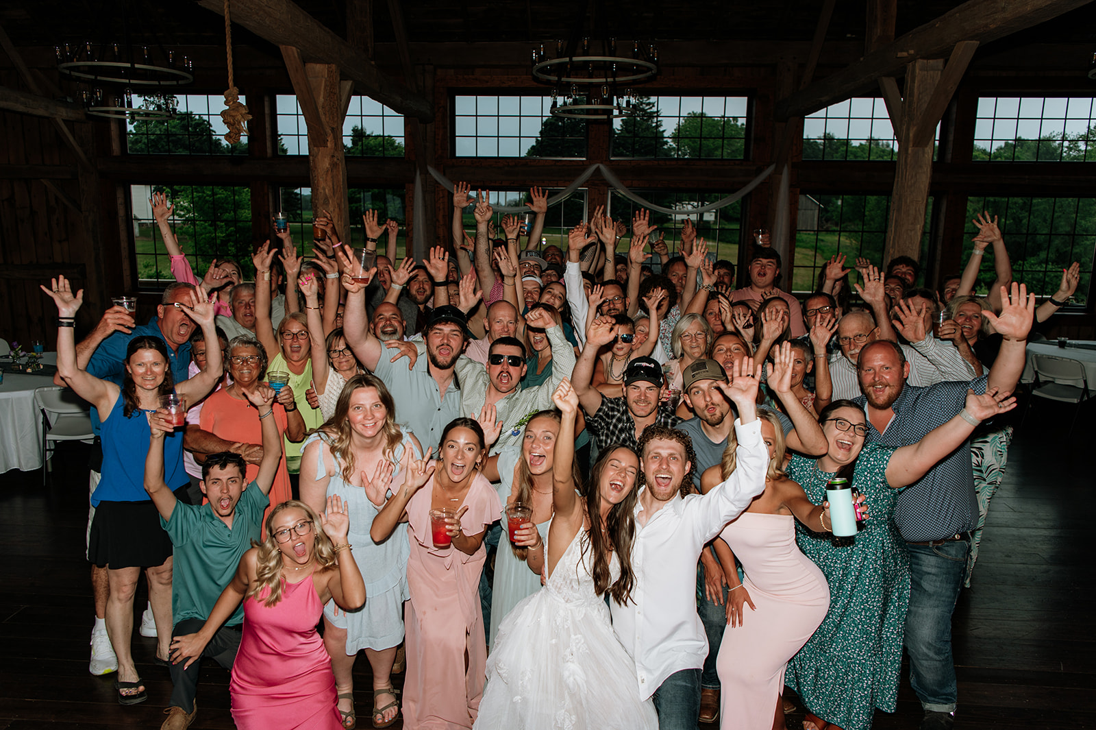 Group photo of the bride, groom, and all their wedding guests inside the barn, cheering and raising their hands together.