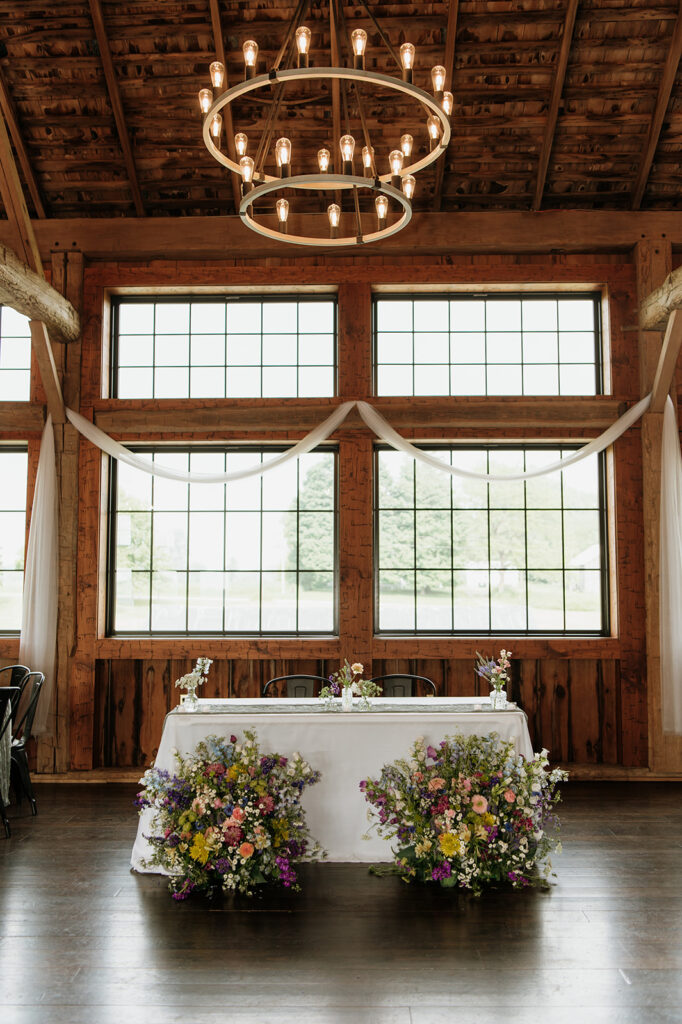 Sweetheart table at 5C Farms with colorful floral arrangements on the floor in front, framed by rustic wood beams and large barn windows.