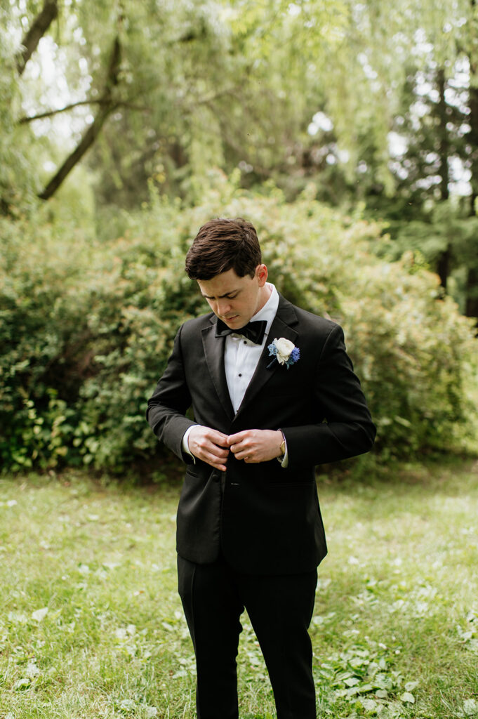 Portrait of George adjusting his black tuxedo jacket with boutonniere, surrounded by greenery for his Notre Dame wedding