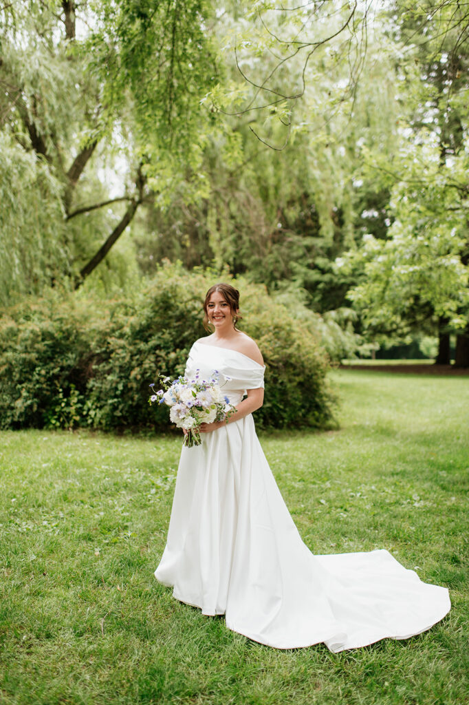 Bride holding her bouquet and smiling in her off-the-shoulder gown, photographed on the green lawn of Saint Mary’s College.