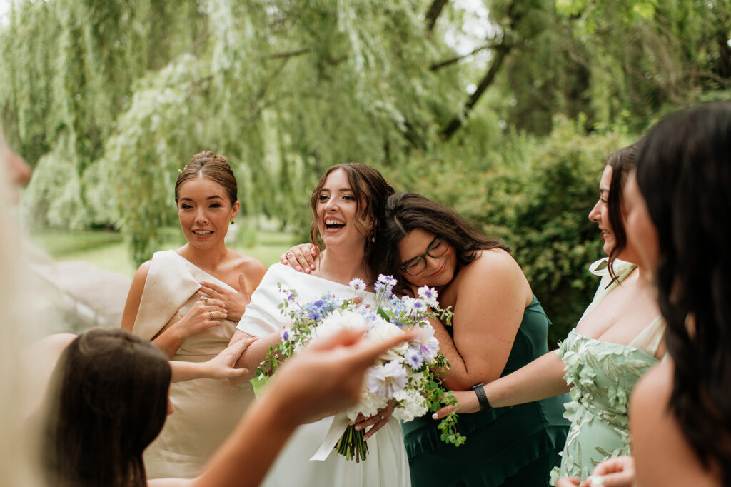 Bride surrounded by bridesmaids, laughing and hugging during candid portraits outdoors.