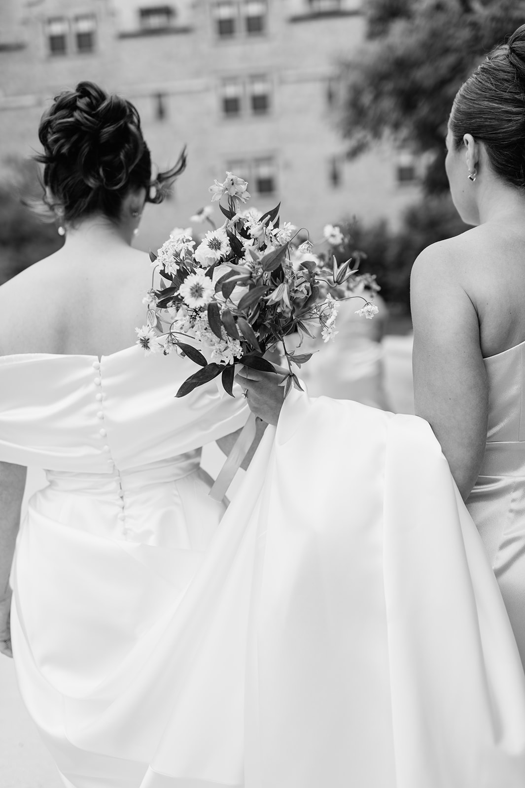 Black and white photo of bride walking with her gown and bouquet, bridesmaid helping to carry her train for her Notre Dame wedding