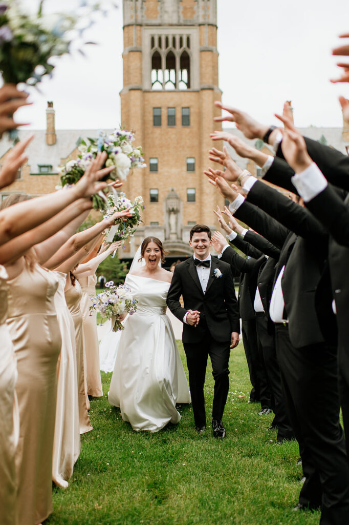 Bride and groom walk hand in hand through a tunnel of bridesmaids and groomsmen raising bouquets and arms outside LeMans Hall at Notre Dame.