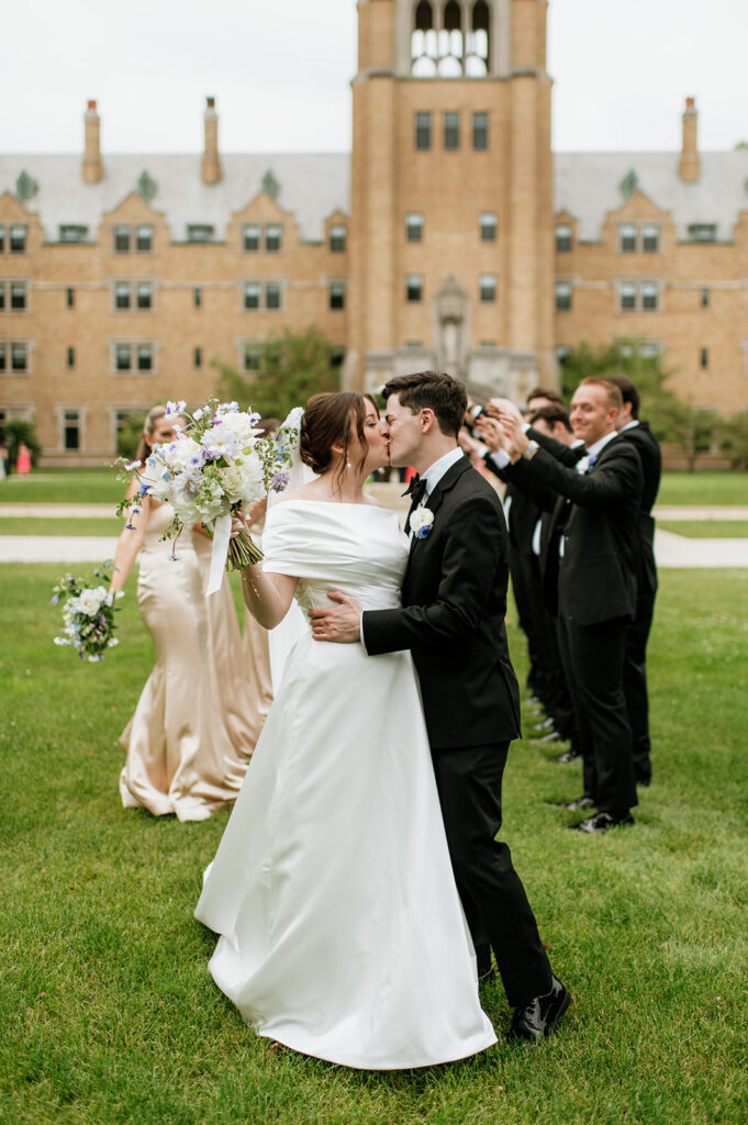 Bride and groom share a celebratory kiss surrounded by their wedding party with LeMans Hall in the background.