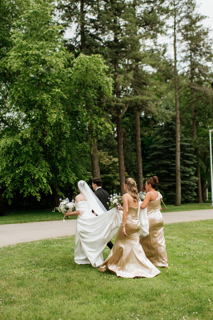 Bridesmaids help carry the bride’s long satin train as she and the groom walk together across the Saint Mary’s College campus.