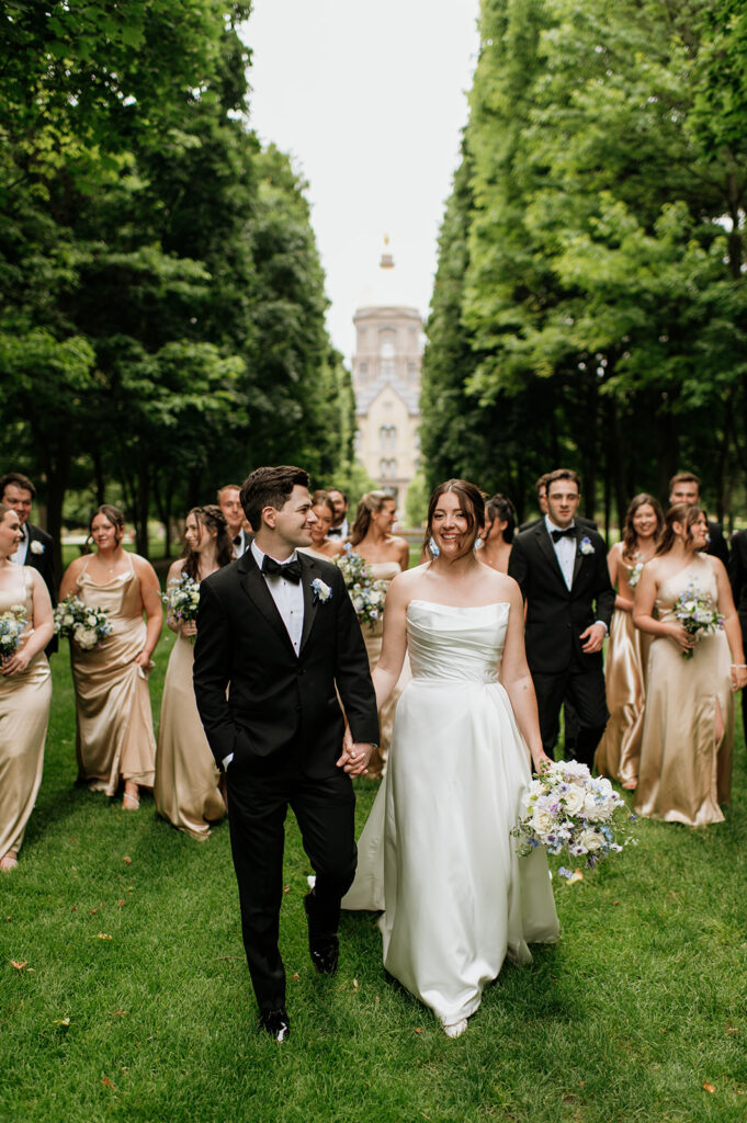 Bride and groom smile at each other while walking hand in hand, surrounded by their bridal party, with the Golden Dome of Notre Dame in the distance.