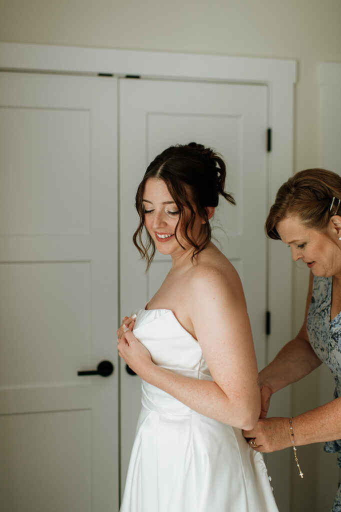 Bride smiling as her mom buttons the back of her gown inside the getting ready space for her Notre Dame wedding