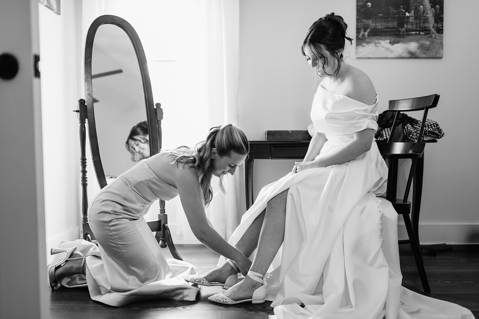 Black and white photo of bridesmaid kneeling to tie the bride’s lace wedding shoes while bride sits in her gown for her Notre Dame wedding