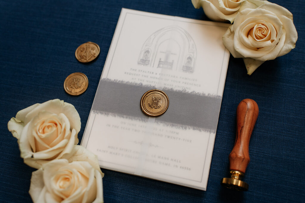 University of Notre Dame wedding invitation with custom gold wax seal on a navy backdrop, surrounded by ivory roses and stamps.