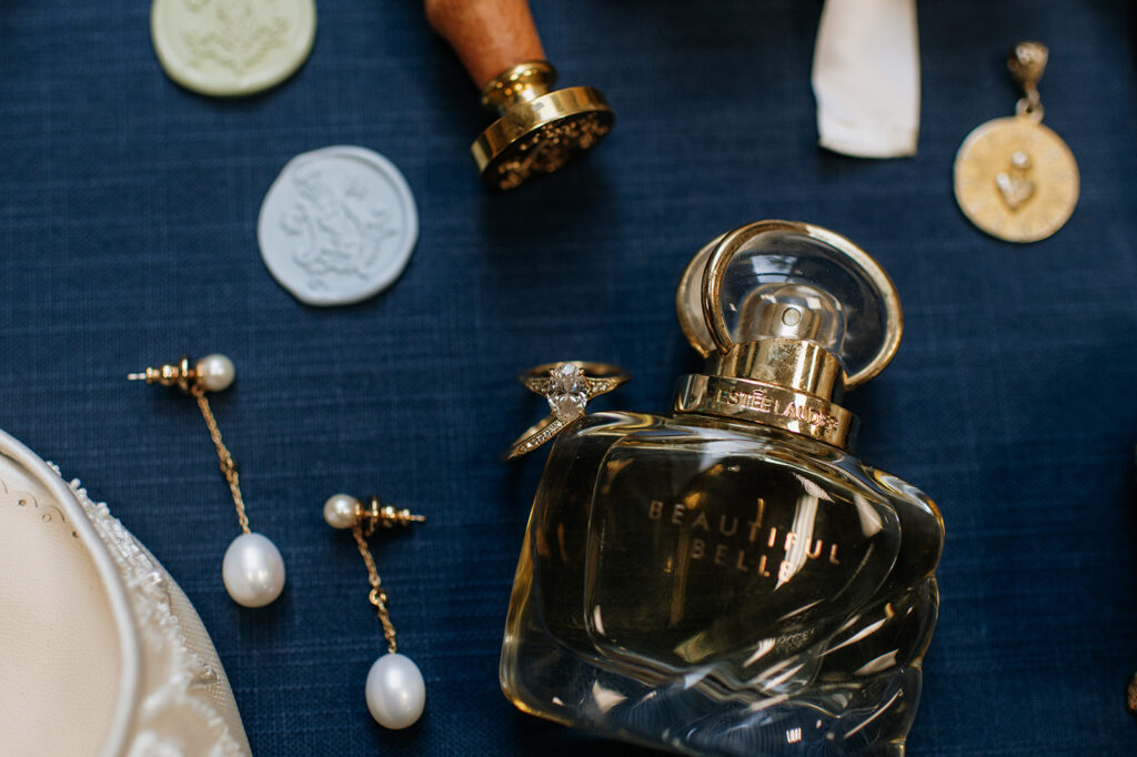 Estée Lauder Beautiful Belle perfume bottle with engagement ring balanced on top, surrounded by pearl earrings and saint pendants.