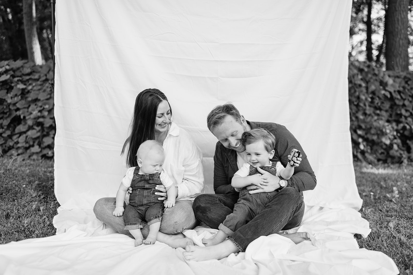 Family of four relaxing barefoot together on the sheet backdrop, smiling at the camera.