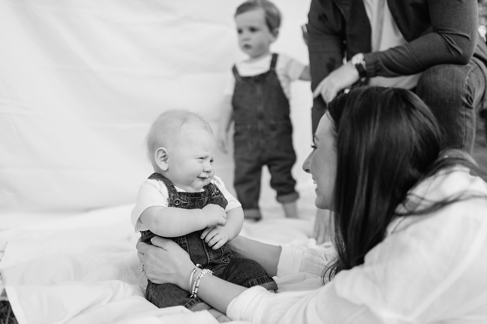 Baby smiling up at mom while lying on a simple sheet backdrop during family photos.