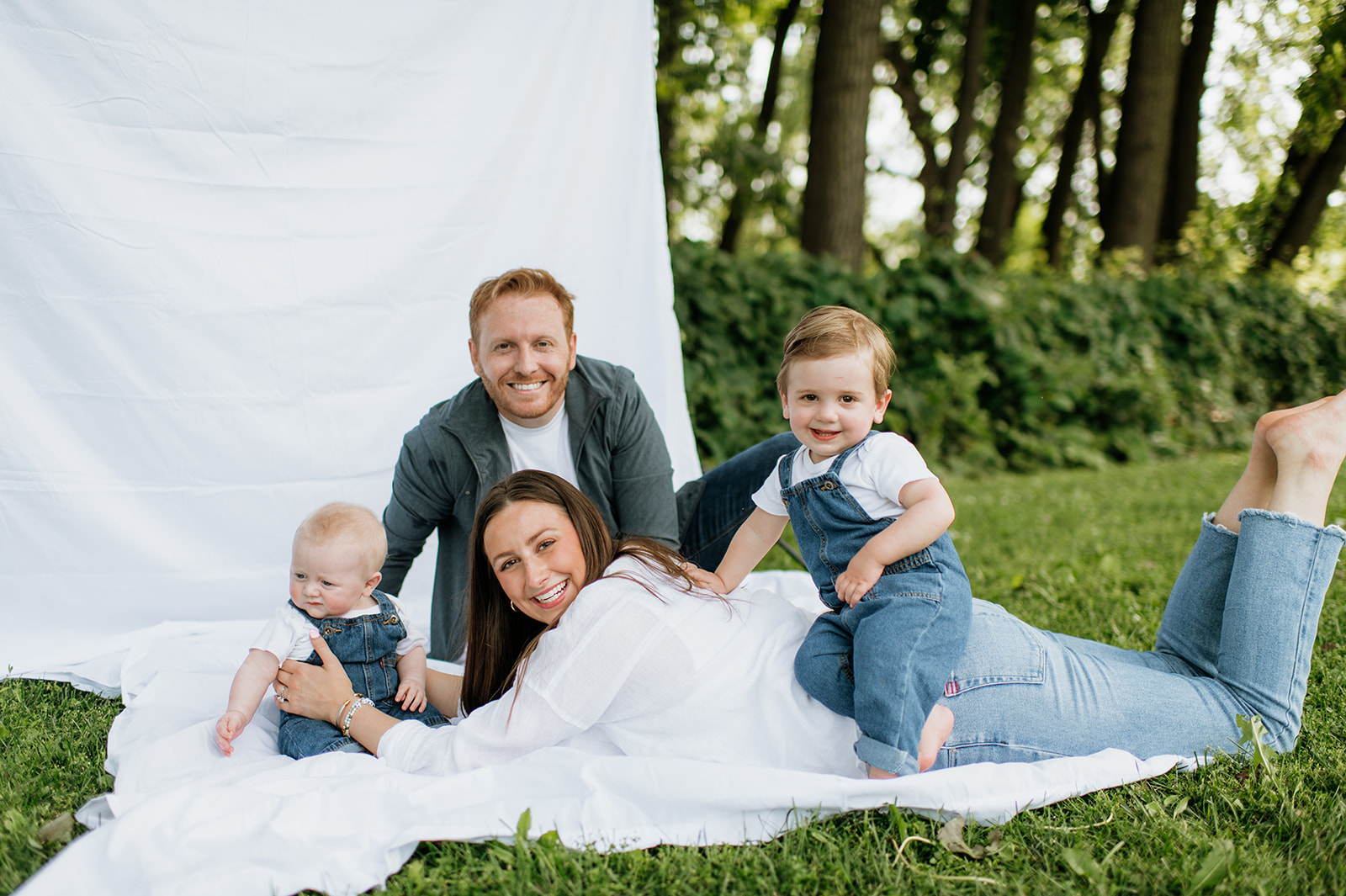 Family of four relaxing barefoot together on the sheet backdrop, smiling at the camera.