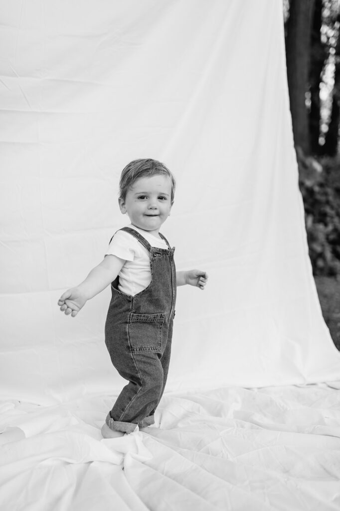 Black and white photo of a toddler walking on a simple white sheet backdrop setup