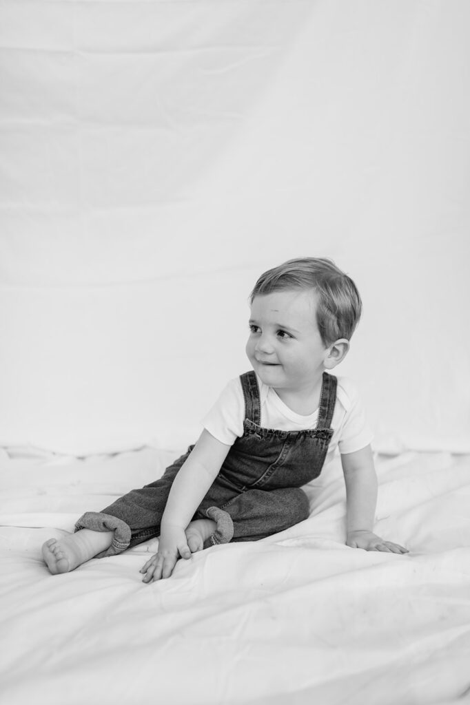 Black and white photo of a toddler boy sitting on a simple white sheet backdrop