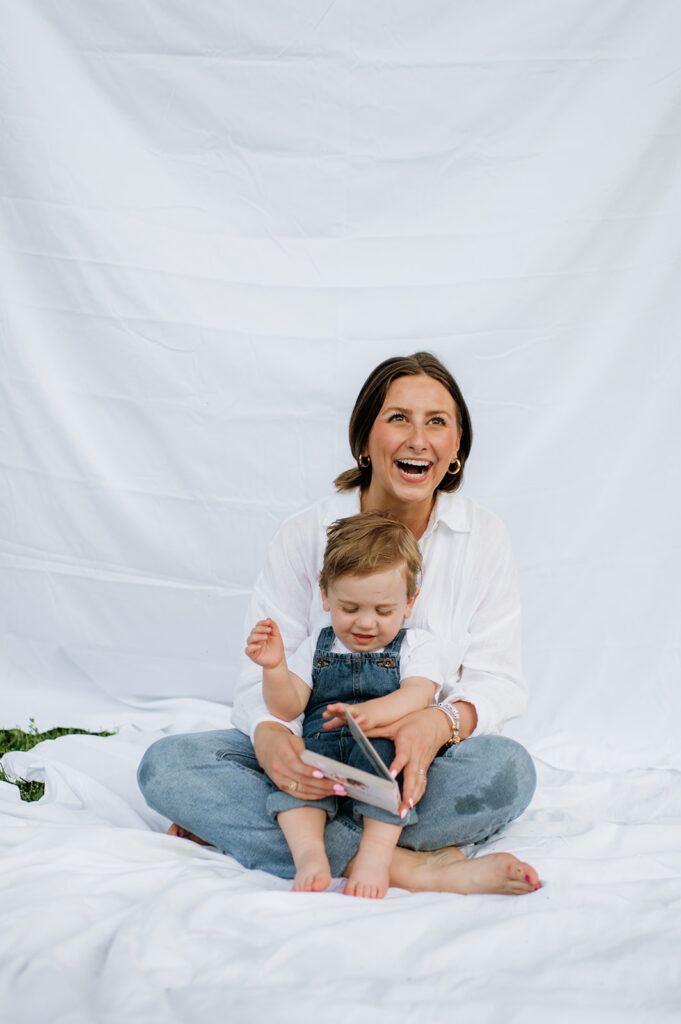 Mom laughing with her toddler on her lap in front of a white sheet backdrop.