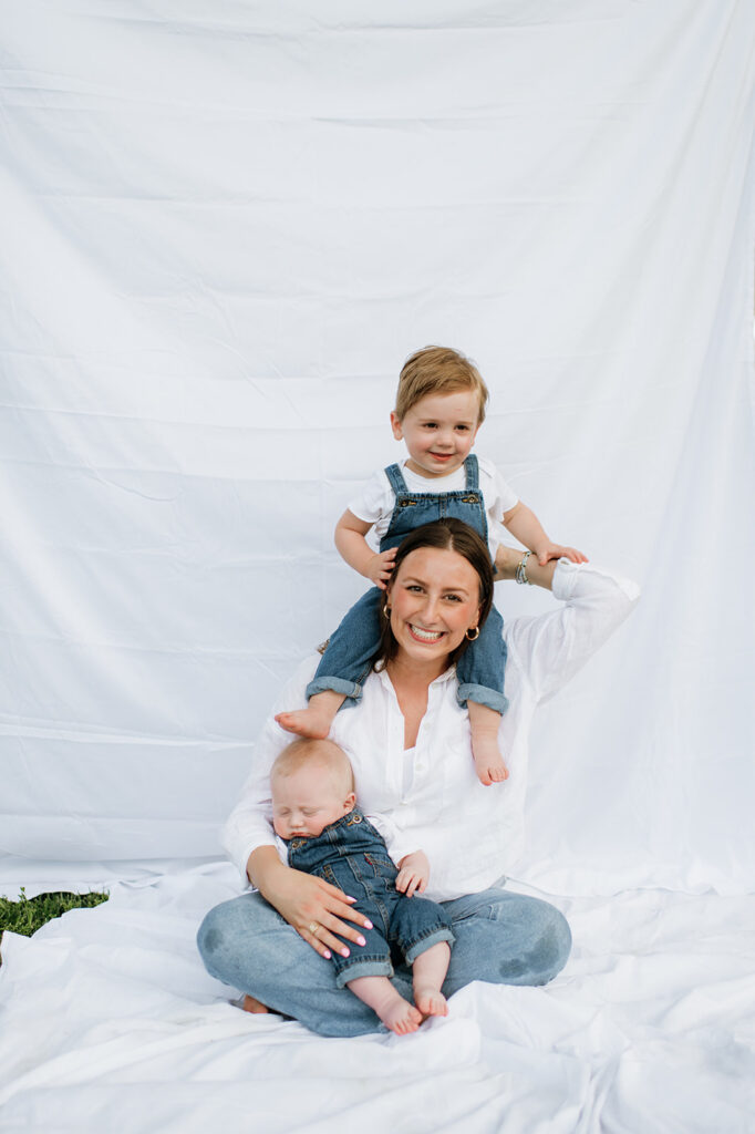 Mom sitting with one child on her shoulders and the other in her lap, both in matching overalls, in front of a simple white sheet.