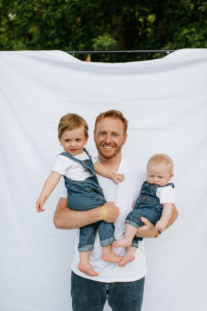 Dad holding his two young sons, one on each arm, in front of a sheet backdrop during their Chicago family photography session.
