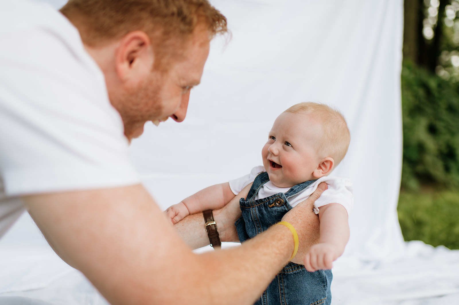 Baby laughing up at dad while sitting on the sheet backdrop, both smiling during family photos.